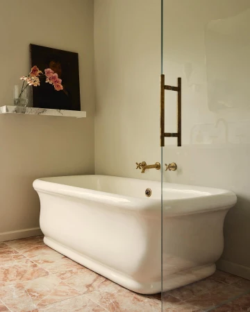 Freestanding white bathtub with gold hardware and glass shower door in a bathroom with patterned tile flooring and neutral-toned walls.