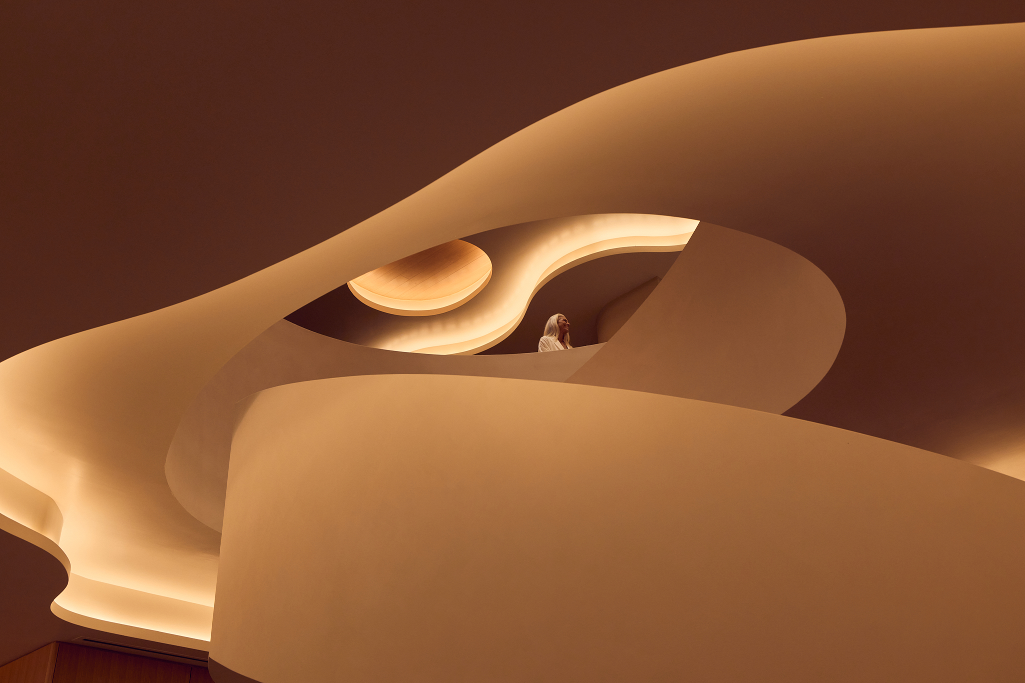 A person with light hair standing on a curving balcony in a minimalist interior with warm, indirect ambient lighting and flowing architecture.