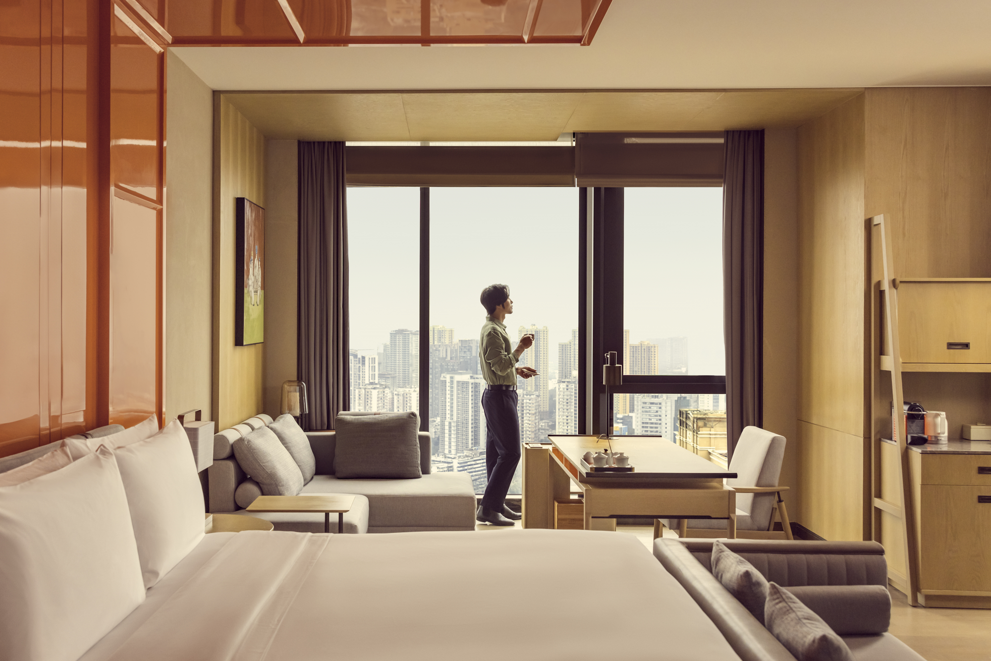 A man in a green shirt and dark trousers stands looking out of a large window in a modern luxury hotel room overlooking a city skyline.