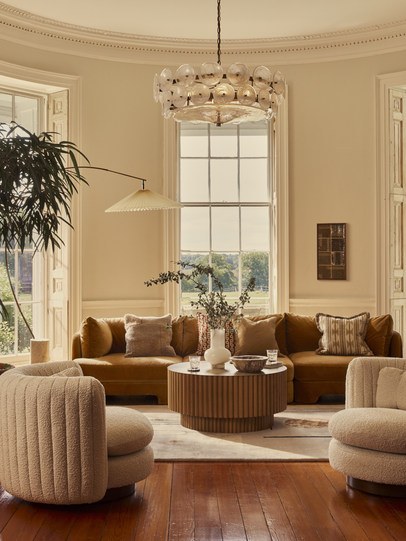 A beige circular room features two curved cream-colored bouclé armchairs and a brown velvet sofa surrounding a round ribbed wooden coffee table.