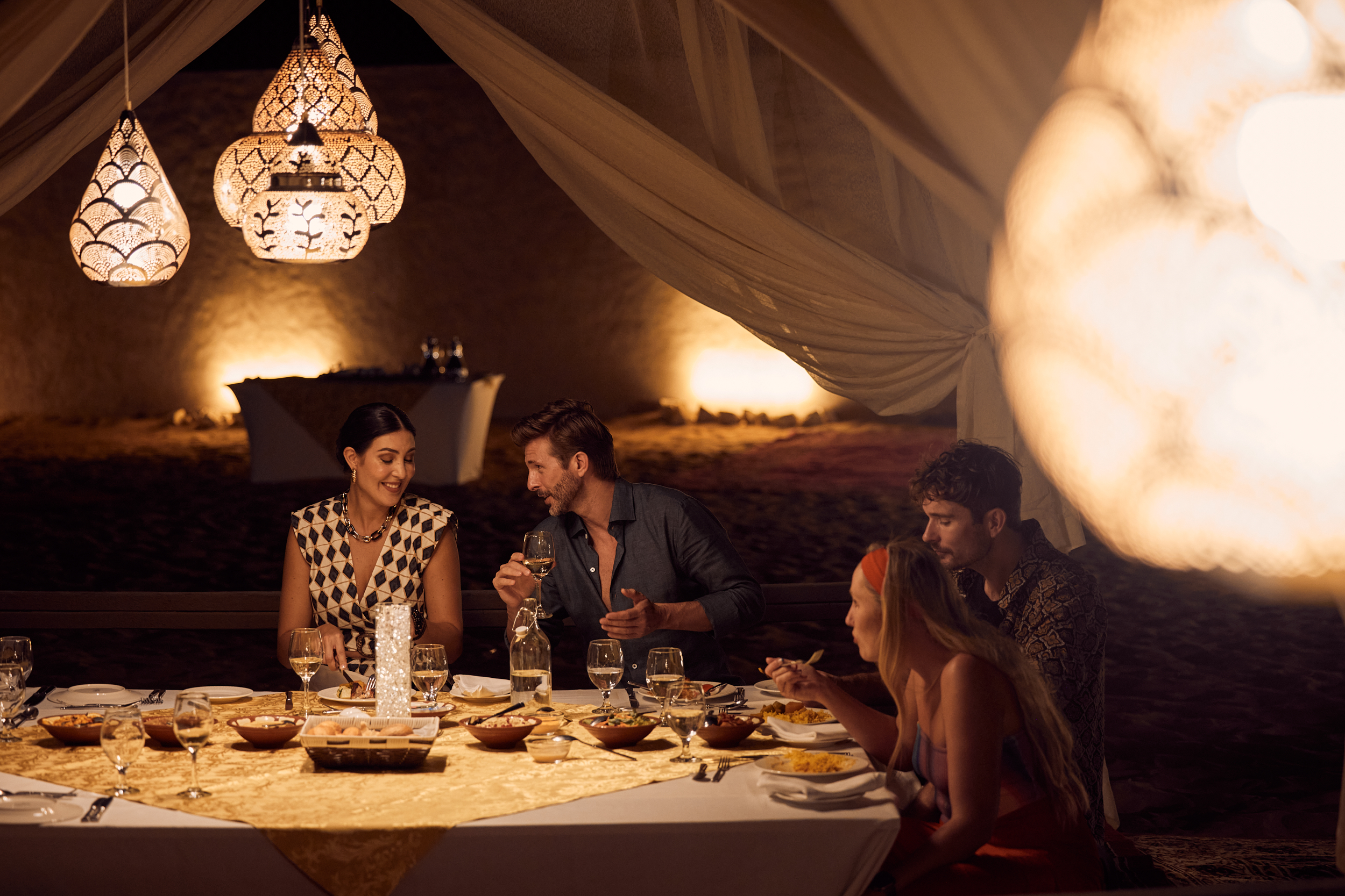 Four people dining at an outdoor table under decorative lamps in a dimly lit, tented space, sharing a meal and conversation.