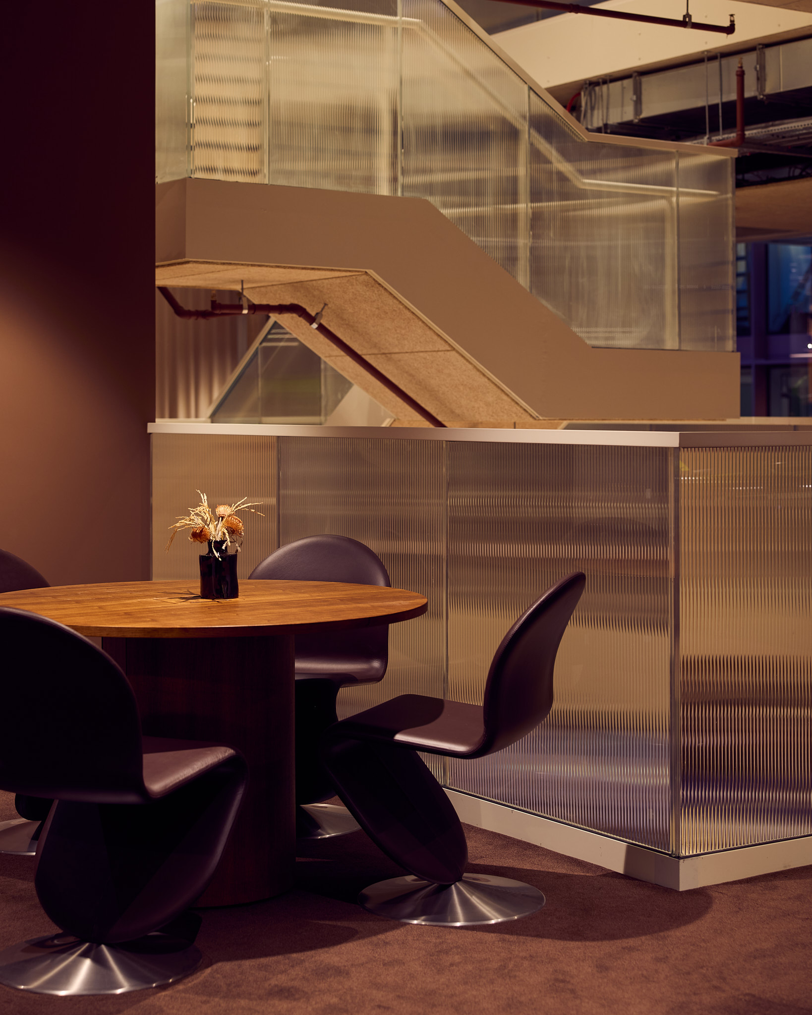 A wooden round table with black swivel chairs in front of a fluted glass partition and a staircase in a modern office space