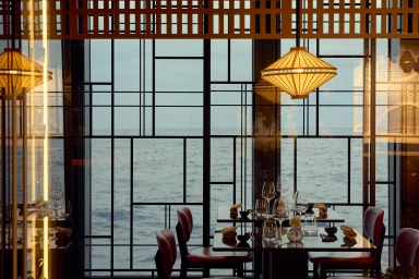 Interior of an upscale restaurant overlooking the ocean through a geometric black metal screen with hanging lantern lamps.