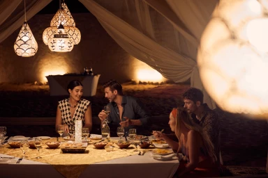 Four people dining around an outdoor table under draped fabric canopies and glowing Moroccan pendant lights photographed by Toby Mitchell