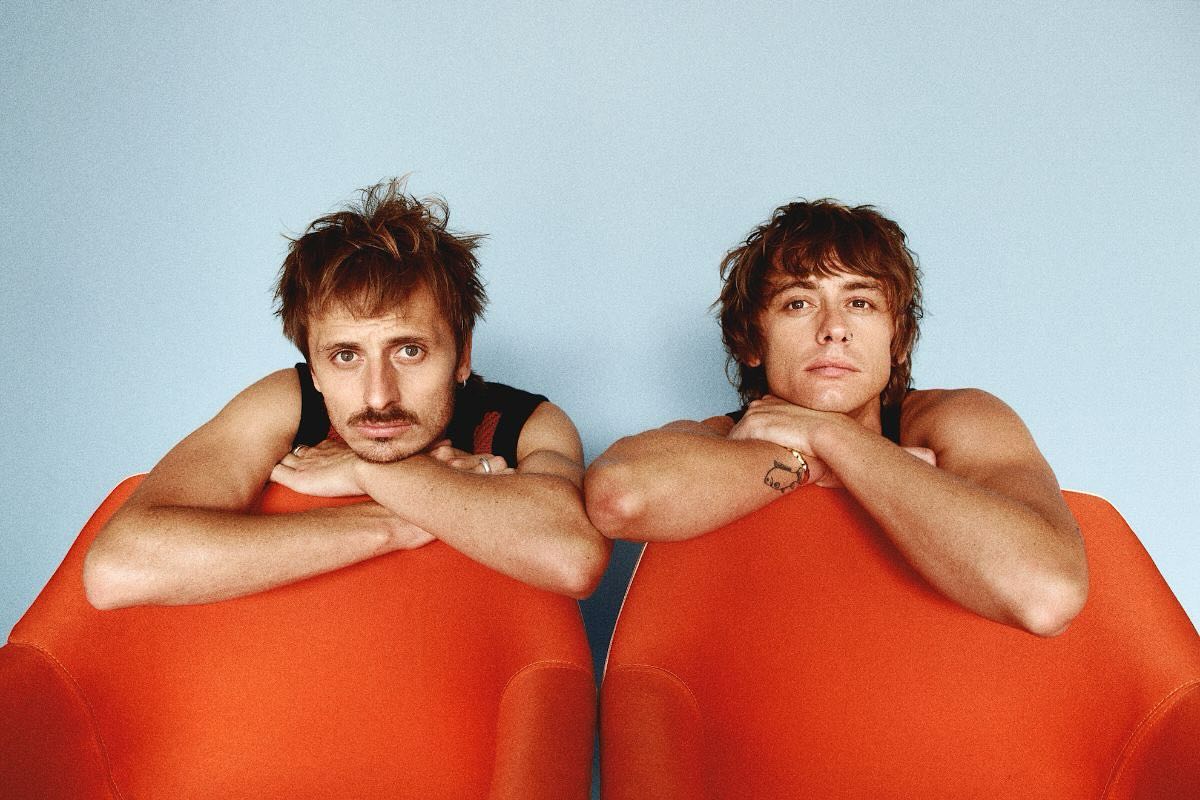 Two adult men, with arms crossed on the tops of orange chairs, pose indoors against a blue background in a medium studio shot.