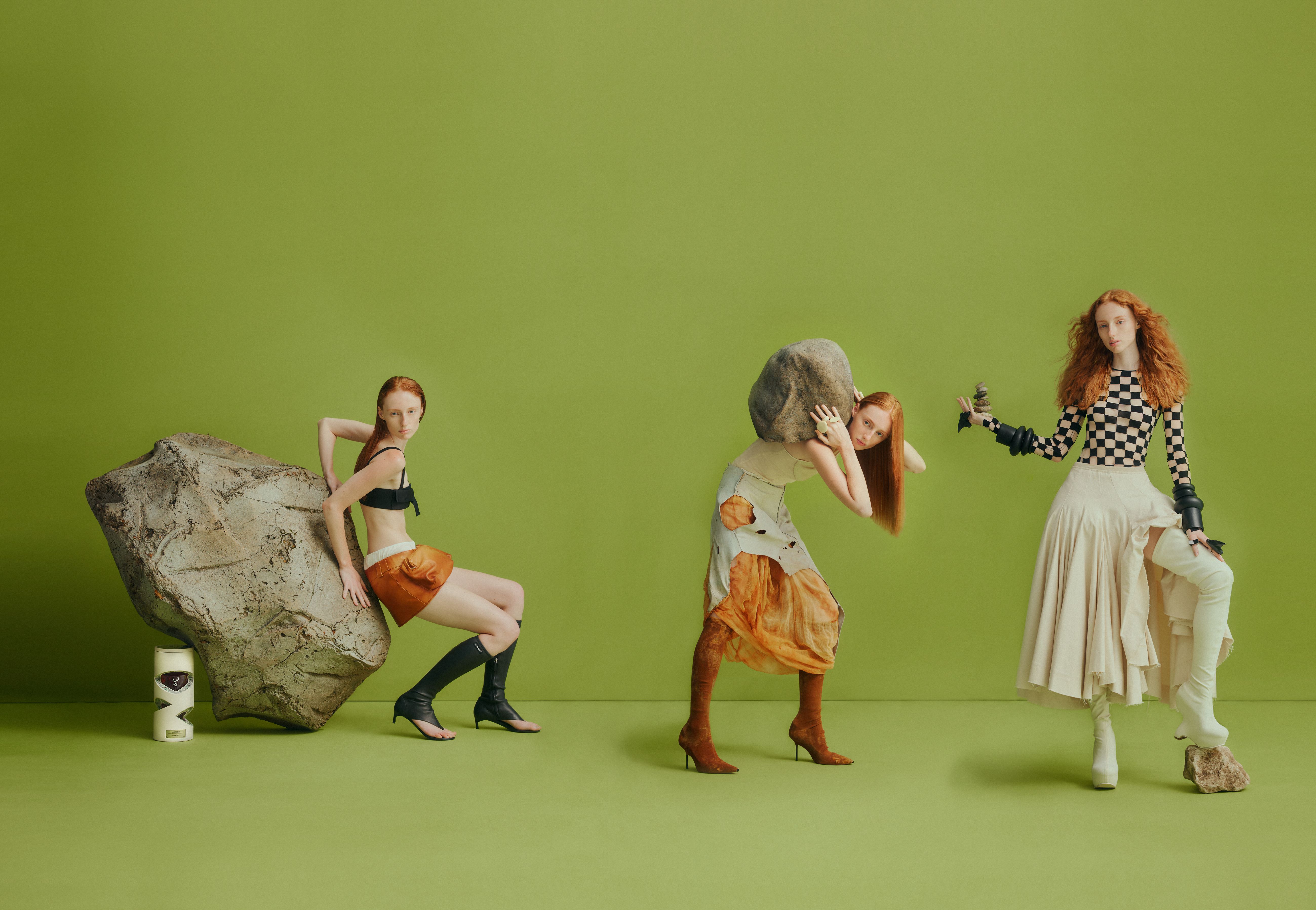 Three red-haired fashion models pose in surreal studio shot with rocks and avant-garde couture clothing on a green backdrop. 