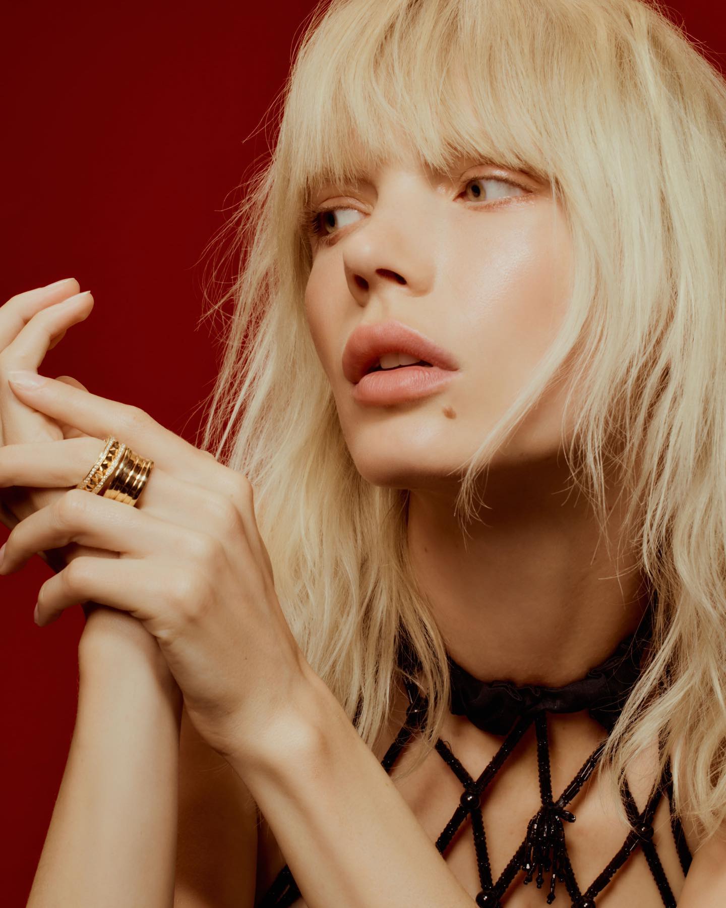 Close-up of a light-blonde adult woman with a mole wearing a black beaded net halter top and gold ring against a red background.