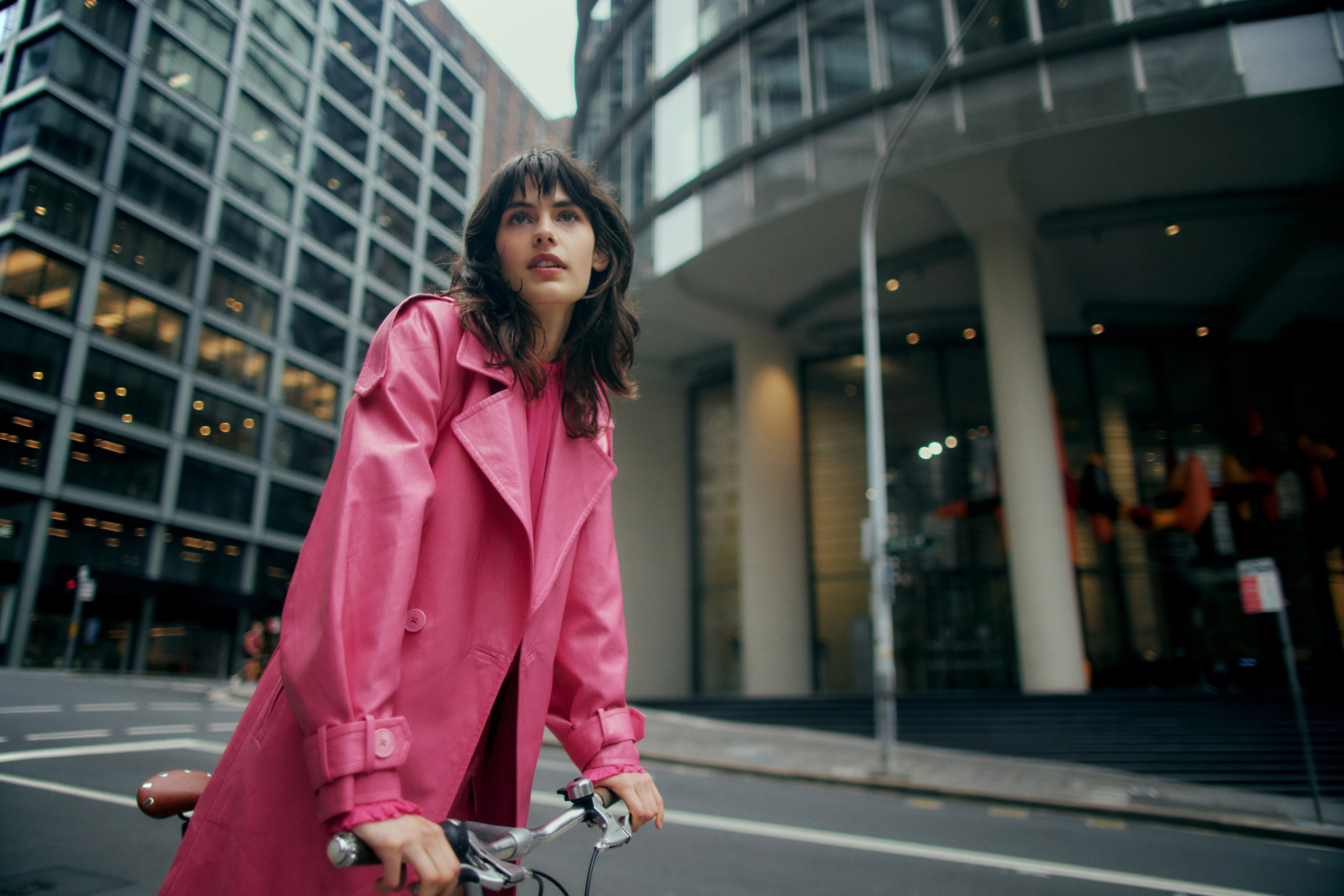 A stylish adult woman in a pink trench coat rides a bicycle on a city street with tall buildings in the background on an overcast day.
