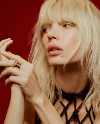 Blonde woman with bangs wearing a wide gold ring and black beaded halter neckwear against a rich red background.