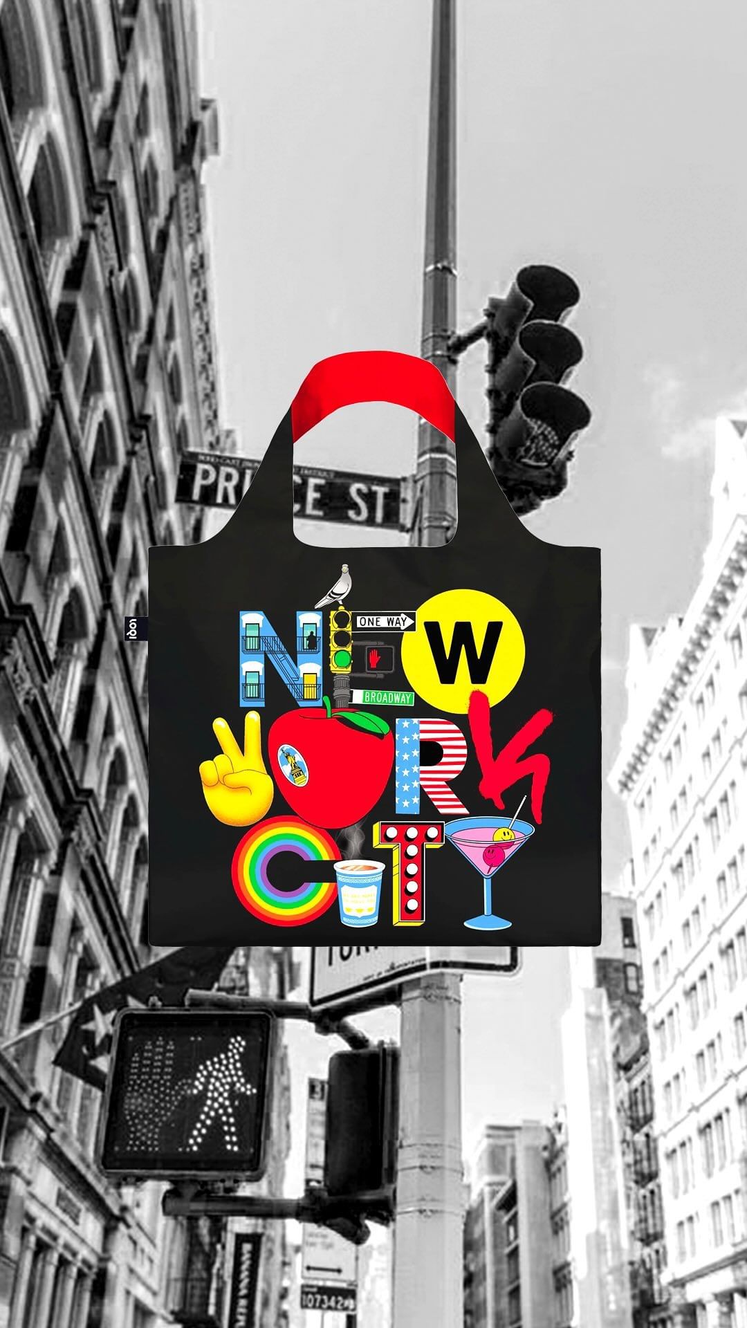 Black tote bag with a New York City design hangs from a traffic light in a black and white urban setting with crosswalk signals.