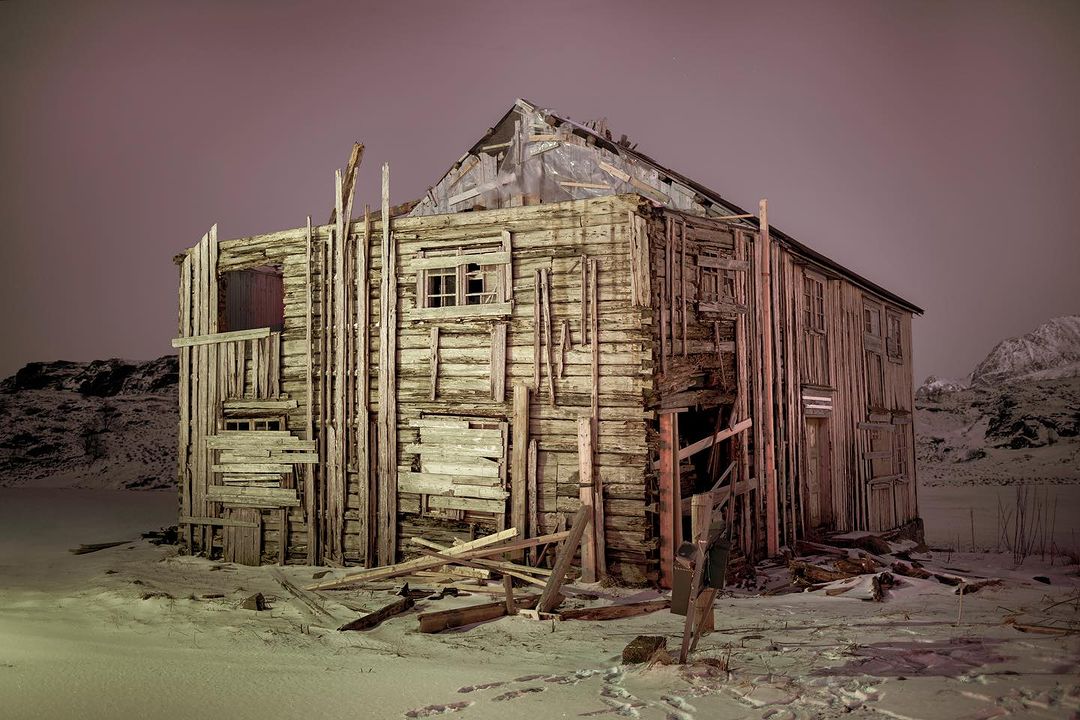 Dilapidated wooden building in a snowy landscape with a pink sky, showcasing decay and a haunting, abandoned atmosphere in nature.