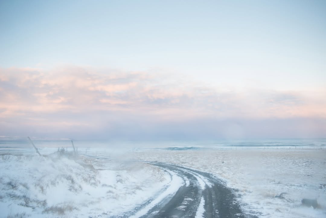 Snowy landscape featuring a road curving through a field covered in snow under a pastel sky with hues of blue, pink, and white.