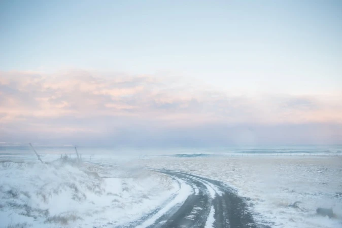 Snowy landscape featuring a road curving through a field covered in snow under a pastel sky with hues of blue, pink, and white.
