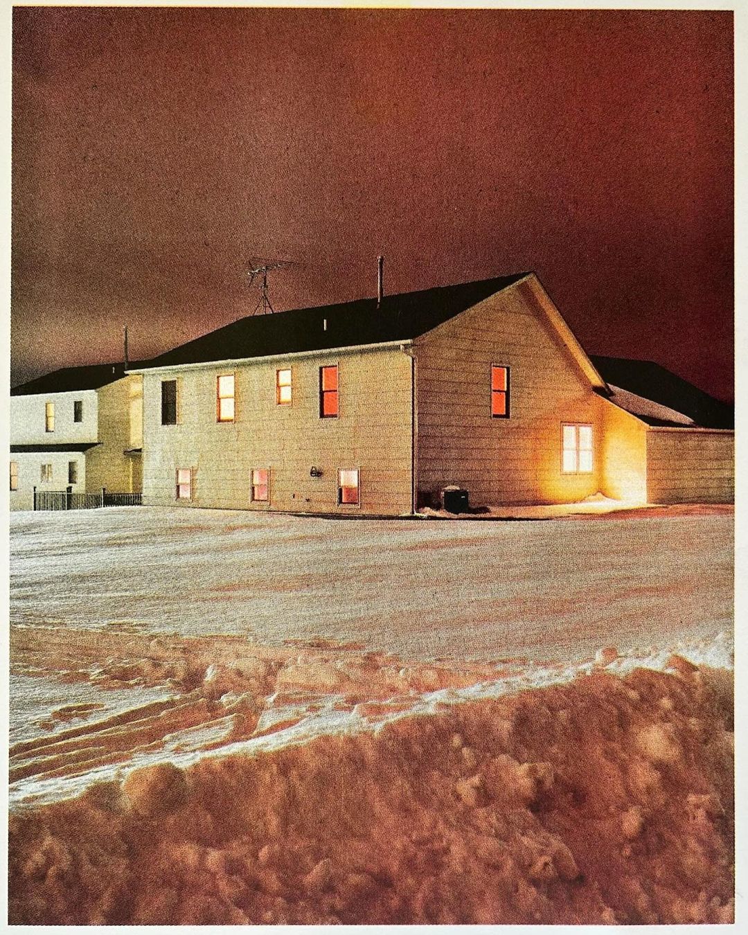 Nighttime scene of a long, off-white apartment complex illuminated by interior lights, set against a dark reddish-brown sky.