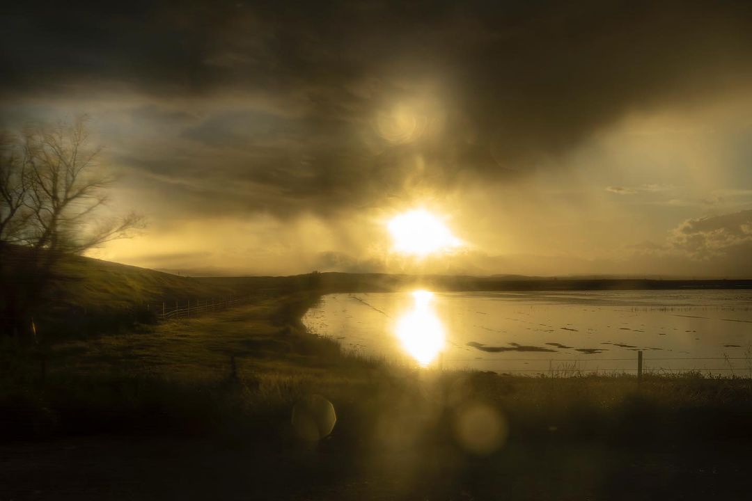 Scenic view of a serene lake reflecting golden sunset light, with grassy shoreline, trees, and a dramatic cloudy sky in the background.