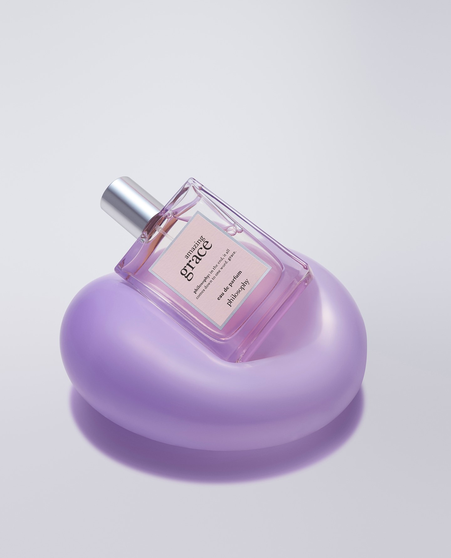 Close-up of a glass Philosophy Amazing Grace perfume bottle on a purple stand, shot in a photo studio against a white backdrop.