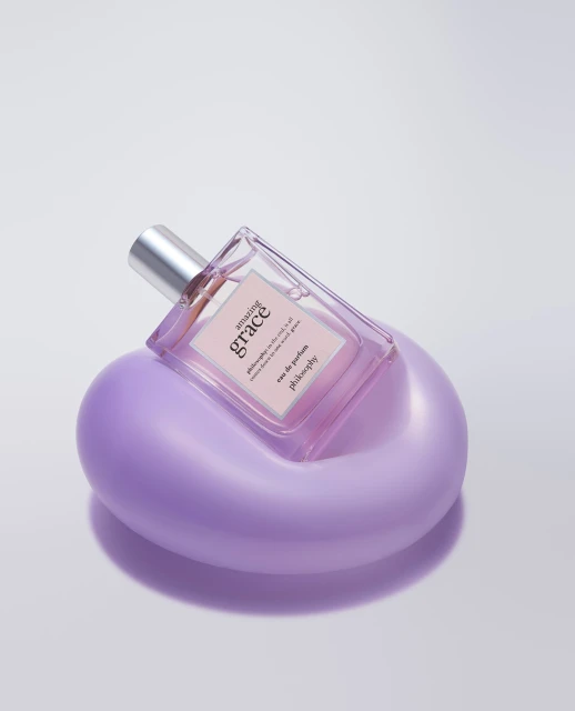 Close-up of a glass Philosophy Amazing Grace perfume bottle on a purple stand, shot in a photo studio against a white backdrop.