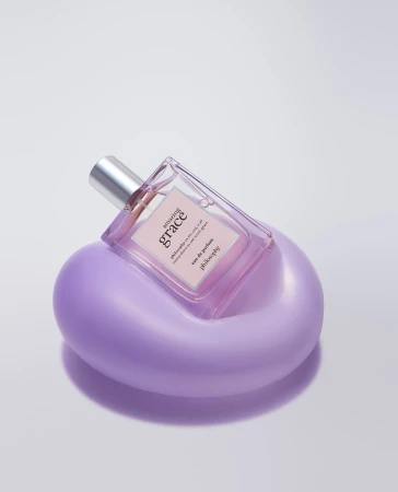Close-up of a glass Philosophy Amazing Grace perfume bottle on a purple stand, shot in a photo studio against a white backdrop.