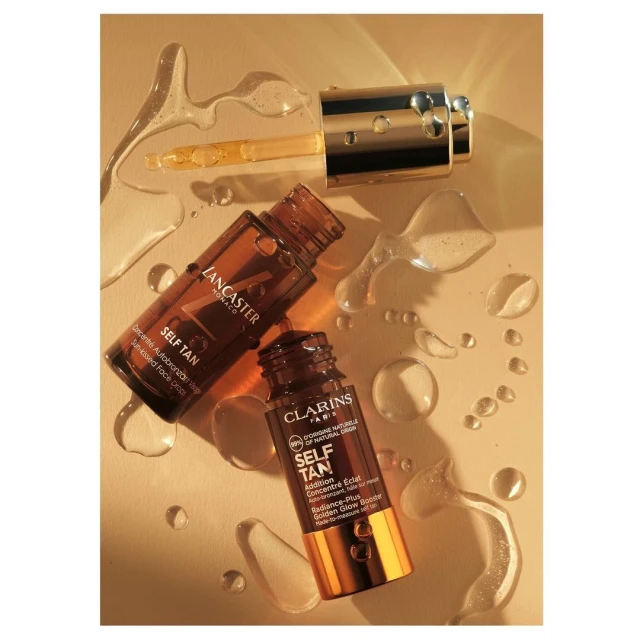 Close-up still life of Lancaster and Clarins self-tanning products, bottles, and a dropper on a light tan background with liquid droplets.