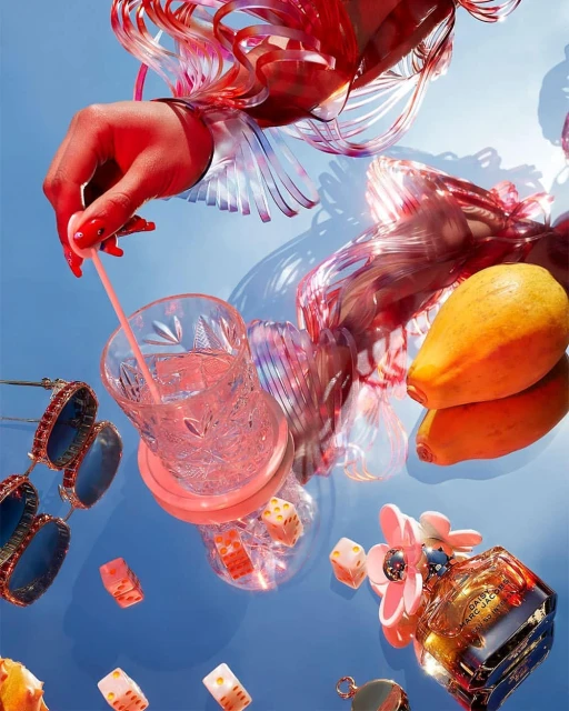 Overhead shot of a hand stirring a drink with clear patterned glass, dice, fruit, sunglasses, and perfume against a blue background.