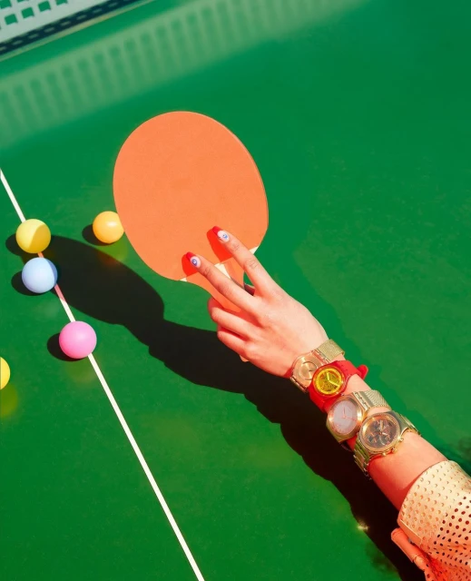 Hand holding orange ping pong paddle on green table with scattered colorful ping pong balls, watches on wrist, bright lighting.