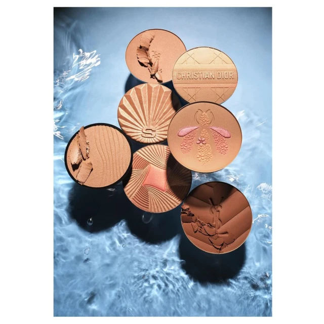 Overhead shot of bronze makeup compacts in a water basin. Compacts display seashell, botanical, and geometric designs in the powder.