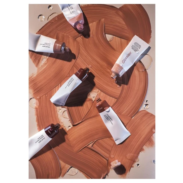 Beauty product flat lay: Glossier Cloud Paint tubes in earth tones against a tan backdrop with smears of product and water droplets.
