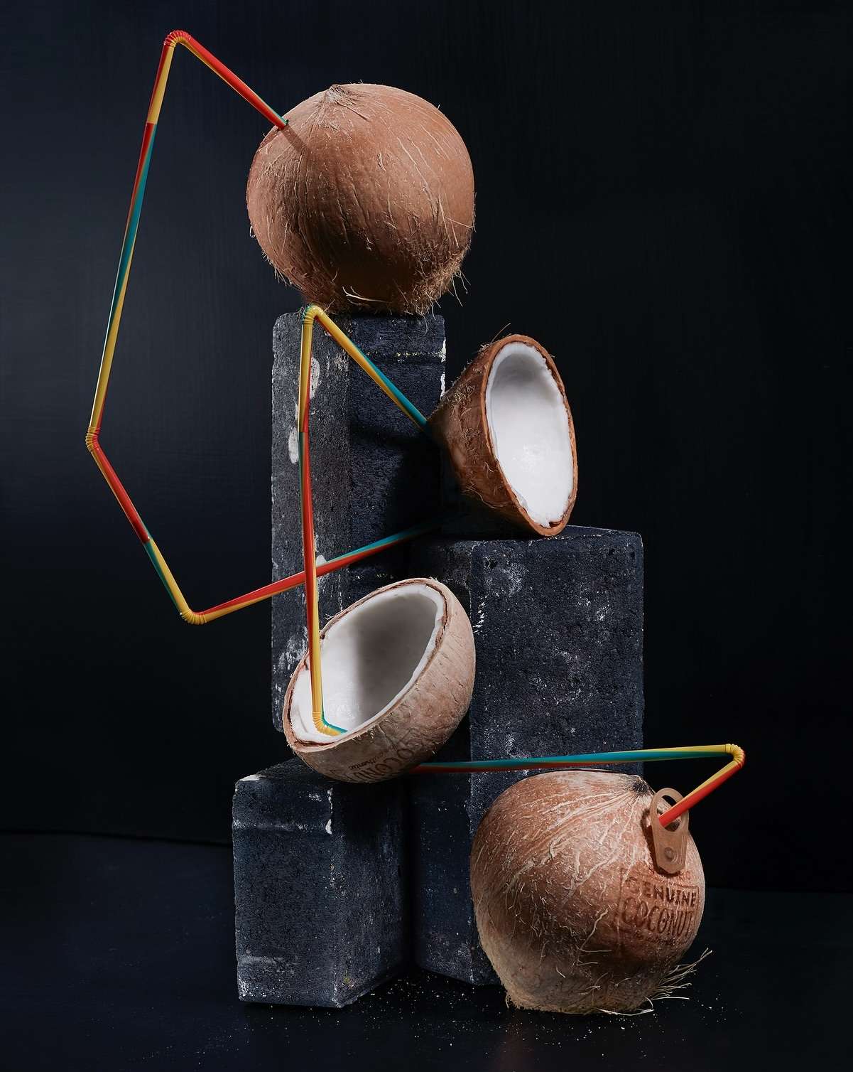 Still life of two coconuts and two coconut halves placed on blocks with a geometric arrangement of colorful drinking straws against black.