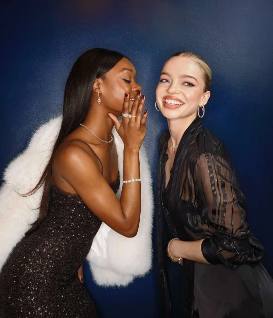 Two women in evening gowns pose in front of a dark blue backdrop; one whispers with a hand near her mouth, both wear jewelry.