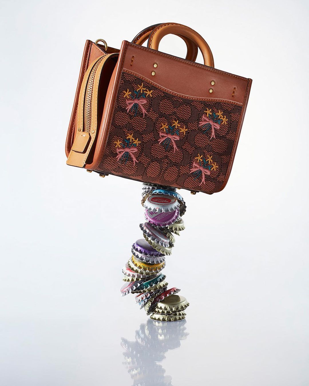 A brown leather purse with floral embroidery sits atop a precarious stack of colorful bottle caps against a white background in a studio shot.