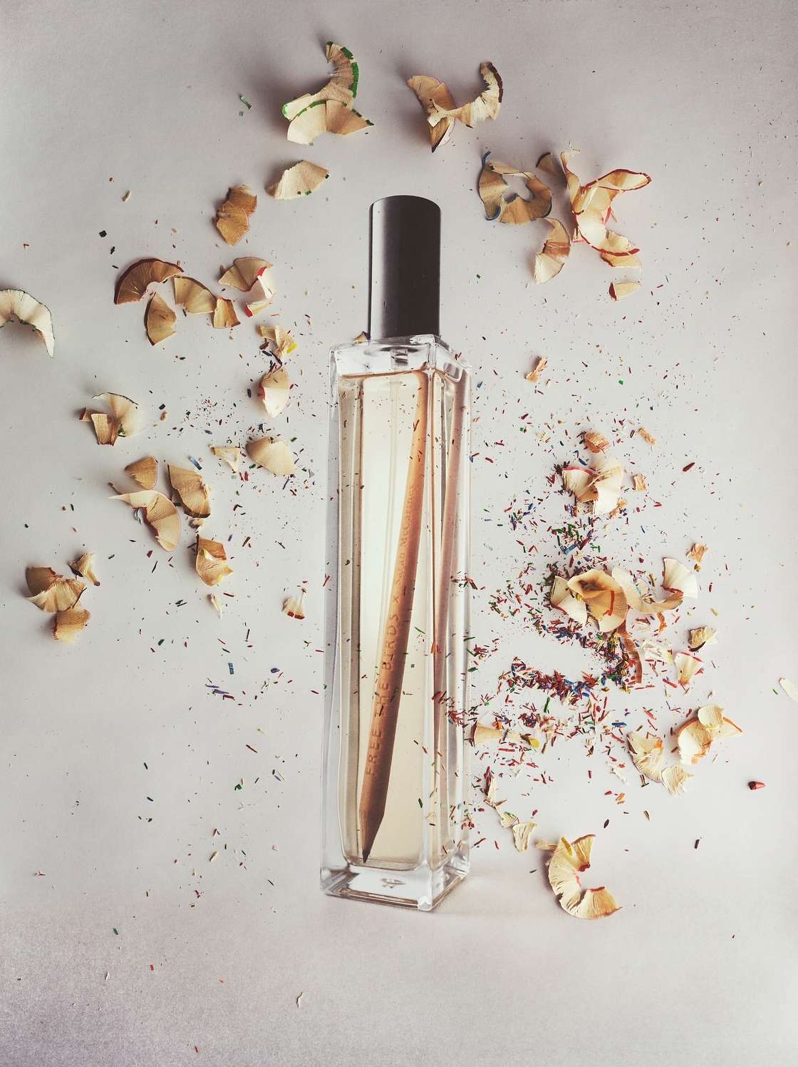 Perfume bottle surrounded by wood shavings and colorful pencil fragments on a white surface in a minimalist studio setting.