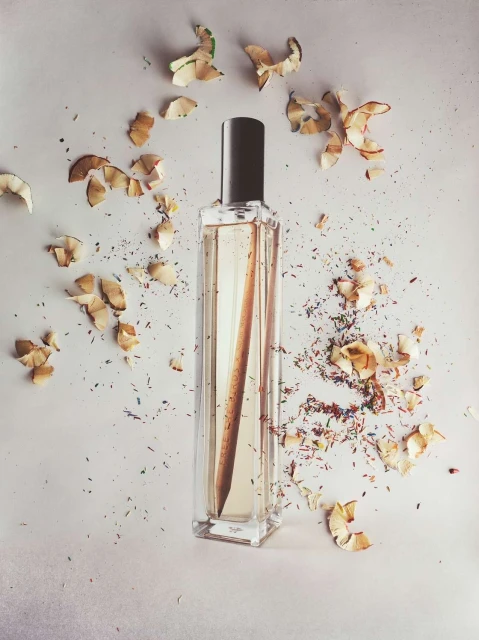 Perfume bottle surrounded by wood shavings and colorful pencil fragments on a white surface in a minimalist studio setting.