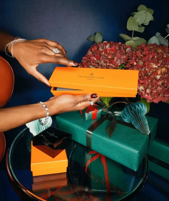 Woman presents jewelry box with ring and bracelet, set on a reflective table with wrapped gifts and floral arrangement, indoors.