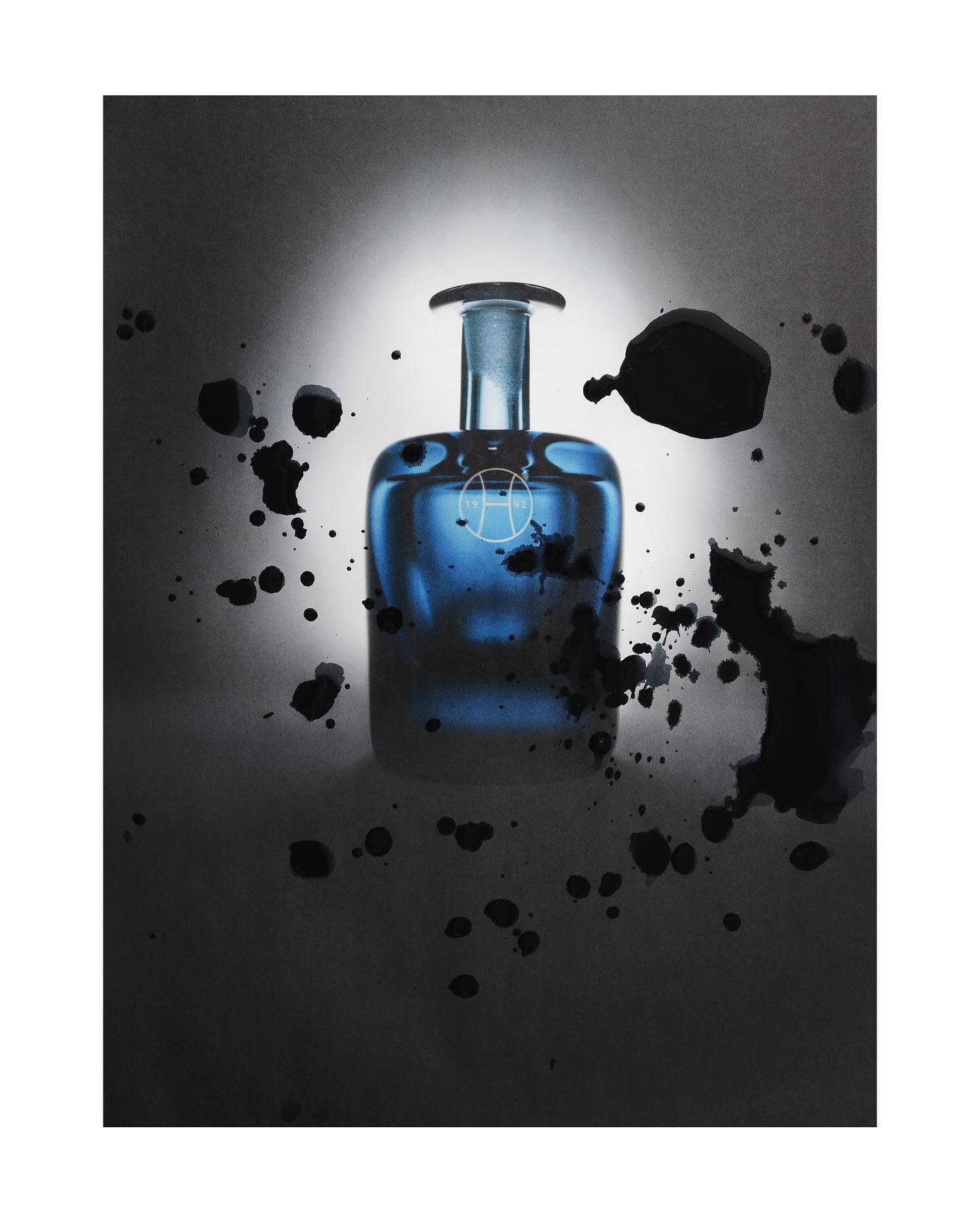 Studio shot of a blue glass perfume bottle with an abstract logo, surrounded by dark ink splashes against a white and grey gradient background.