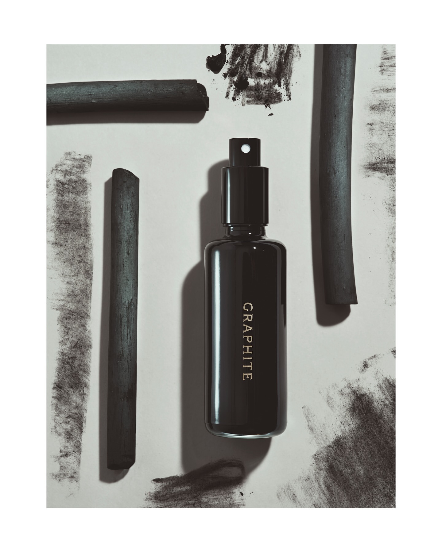 Still life shot of a dark grey perfume bottle with the text "GRAPHITE" and charcoal sticks on a grey and white surface.