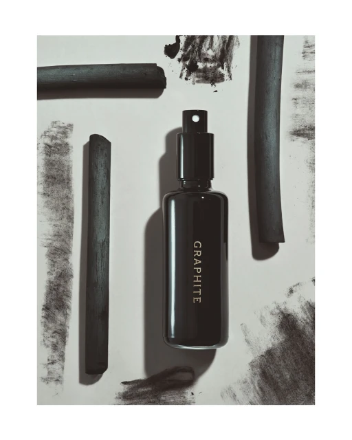 Still life shot of a dark grey perfume bottle with the text "GRAPHITE" and charcoal sticks on a grey and white surface.