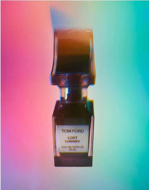 Close-up of a Tom Ford Lost Cherry perfume bottle with a brown cap against a gradient pink, blue, and green background.