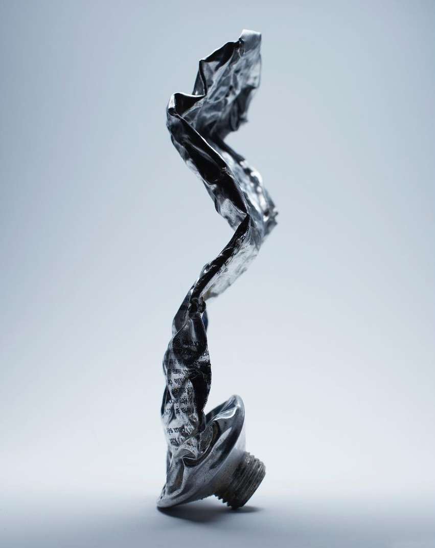Close-up of a shiny, crumpled metal tube standing upright against a gradient light blue background in a studio setting.