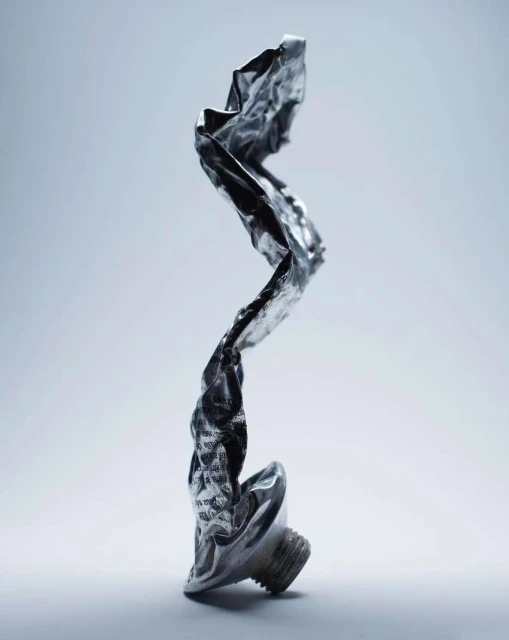Close-up of a shiny, crumpled metal tube standing upright against a gradient light blue background in a studio setting.