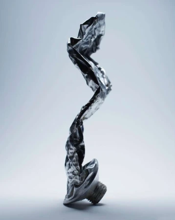 Close-up of a shiny, crumpled metal tube standing upright against a gradient light blue background in a studio setting.