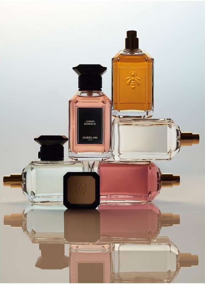 Guerlain perfume bottles and a bronzer compact are stacked on a reflective surface in a beauty still life with white gradient background.