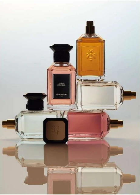Guerlain perfume bottles and a bronzer compact are stacked on a reflective surface in a beauty still life with white gradient background.