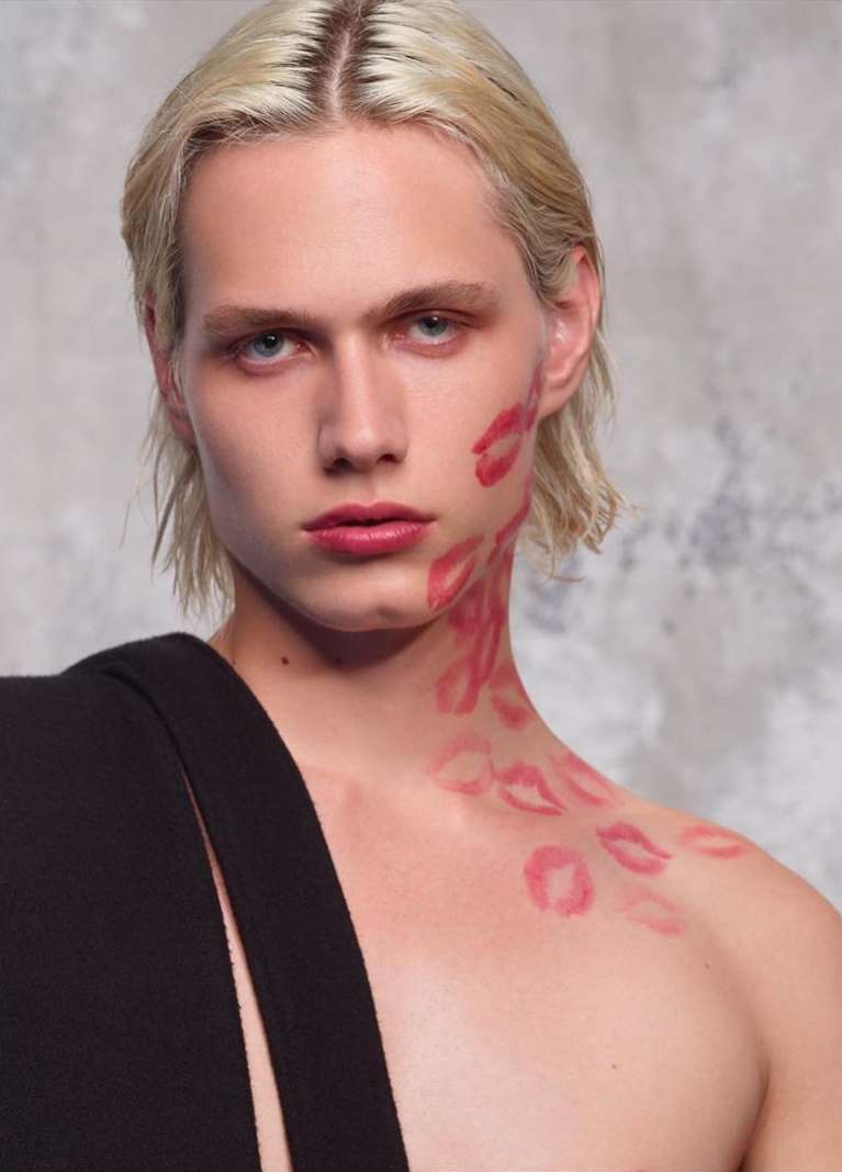 Adult male fashion model with painted lipstick marks on face and neck posing in front of gray background; head and shoulders shot.