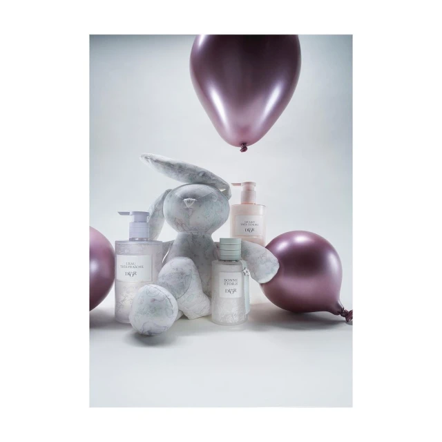 Still life of a plush rabbit toy surrounded by skincare bottles and pink balloons against a soft, neutral studio backdrop. Beauty product focus.