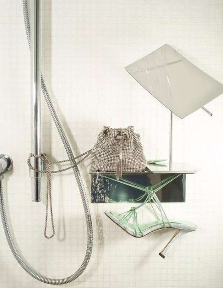 Fashion still life featuring a silver bucket bag, green heeled sandals, and chrome shower fixtures against a white tiled wall.