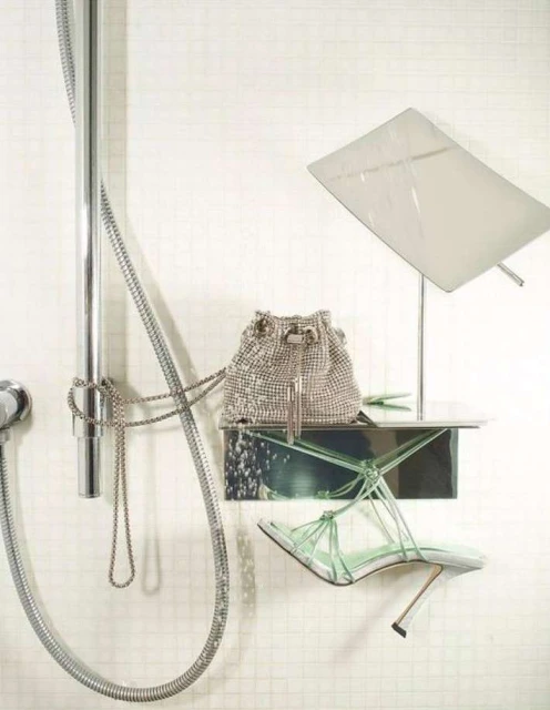 Fashion still life featuring a silver bucket bag, green heeled sandals, and chrome shower fixtures against a white tiled wall.