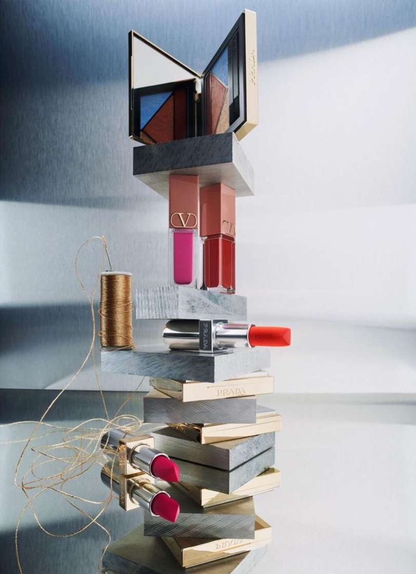 Cosmetics still life featuring lipsticks, lip glosses, and eyeshadow palettes stacked on blocks of gray and gold.