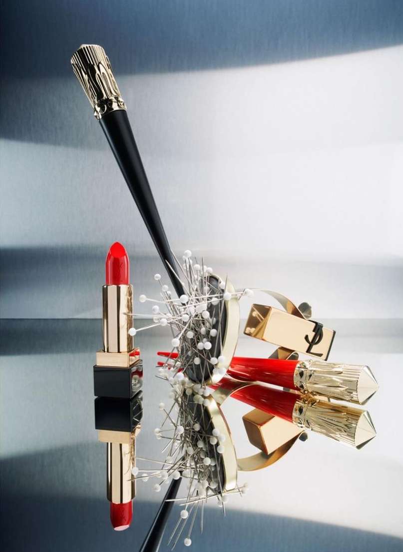 Still life shot of YSL lipstick in red and gold packaging, arranged with a makeup brush and decorative pins on a reflective surface.