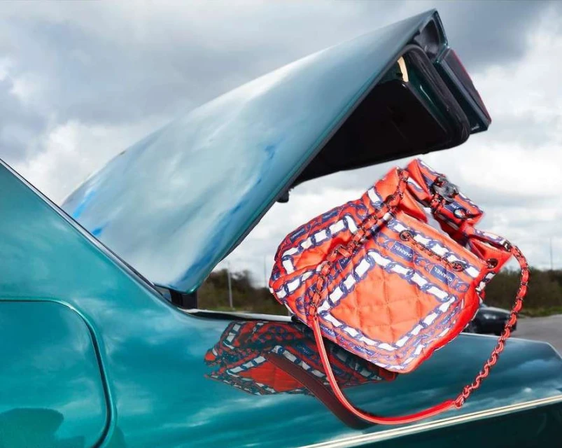 Orange drawstring bucket bag resting on the open trunk of a turquoise vintage car, against a cloudy sky backdrop.