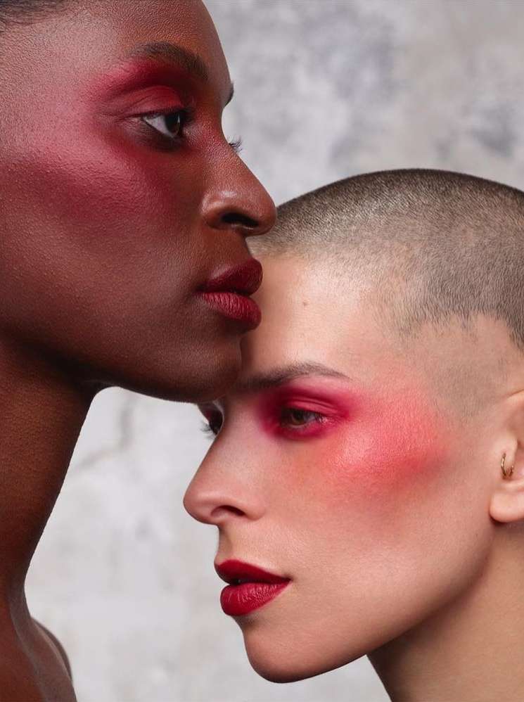Two women with matching red makeup pose in a beauty shot. The woman in front has a shaved head and fair skin. Beauty concept.